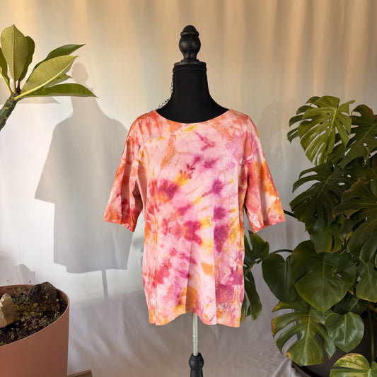 Mannequin wearing a tie-dye shirt with pink and orange patterns, surrounded by plants.